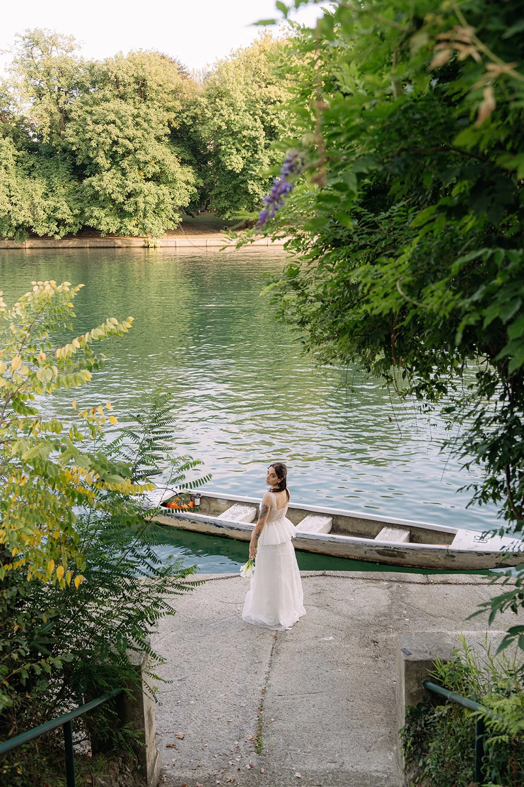 A woman in a wedding dress holding flowers standing on a concrete path near a boat by a lake, surrounded by green trees.