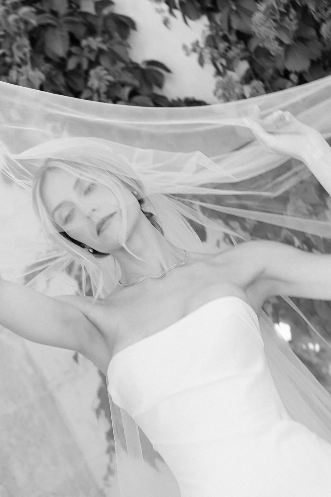 A woman in a strapless white dress lying underneath a sheer fabric with her eyes closed.