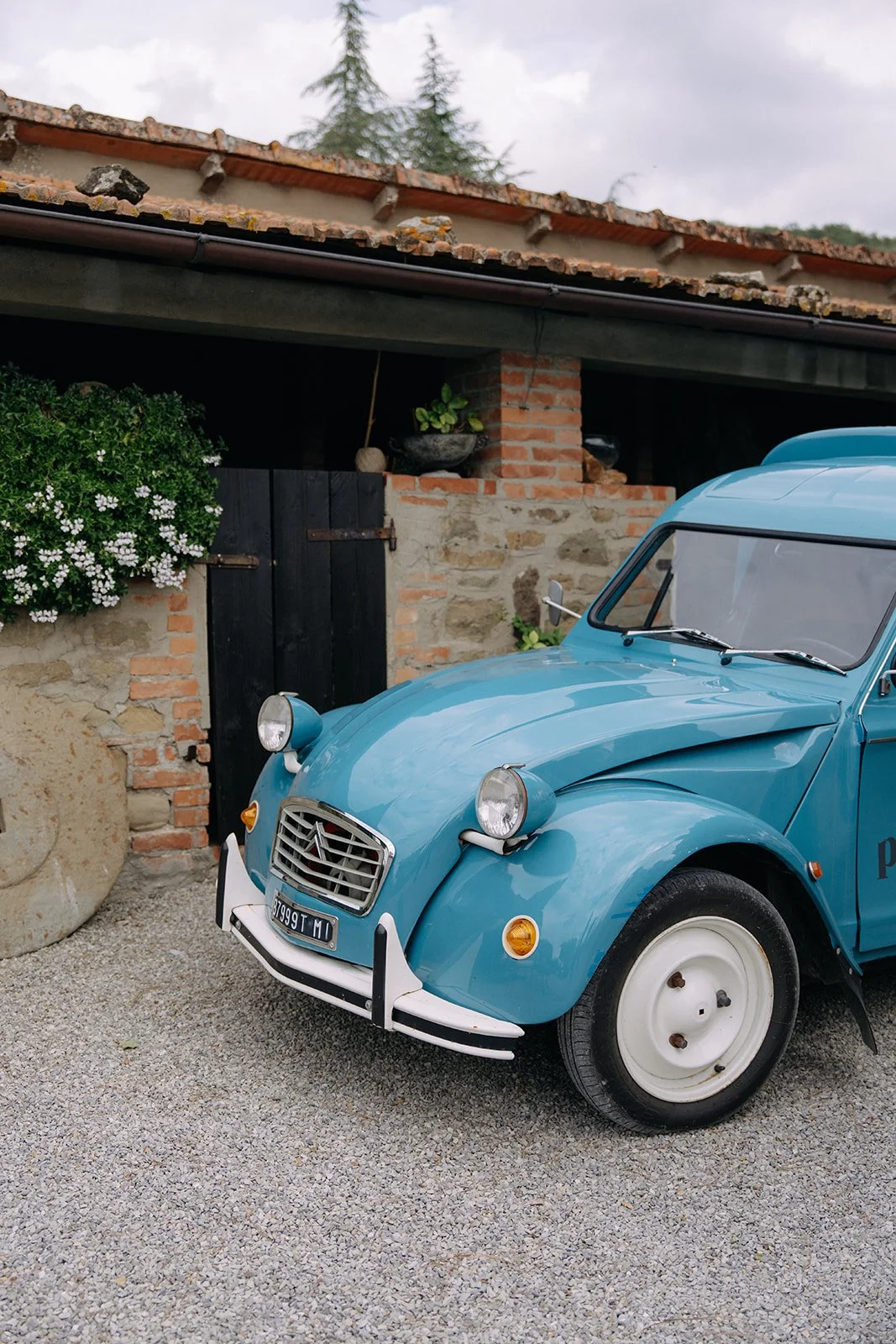 A vintage blue Citroën 2CV car parked on gravel in front of a rustic brick and stone building with a black door, surrounded by greenery and flowering plants.