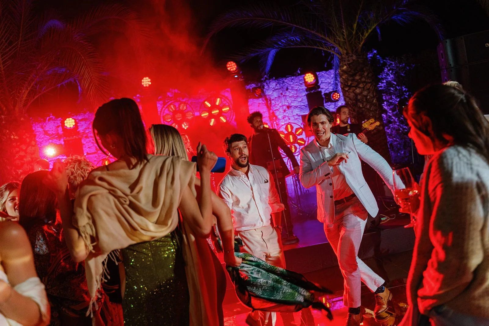 People dancing and singing at a lively party with colorful lights and a live band playing on stage.