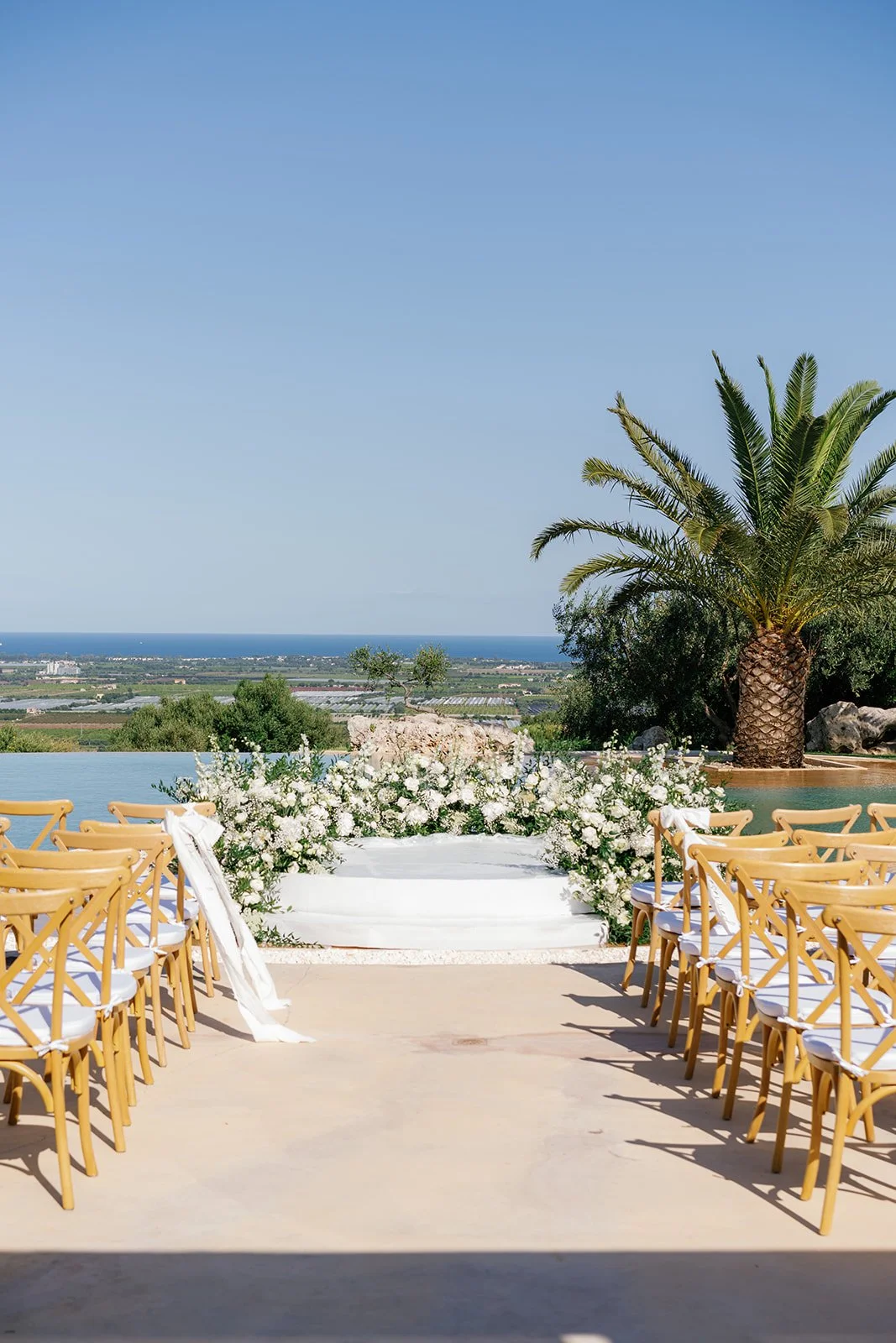 Wedding ceremony setup with chairs decorated with white ribbons, white floral arrangements, and a backdrop overlooking a scenic landscape with trees, a body of water, and a clear blue sky.