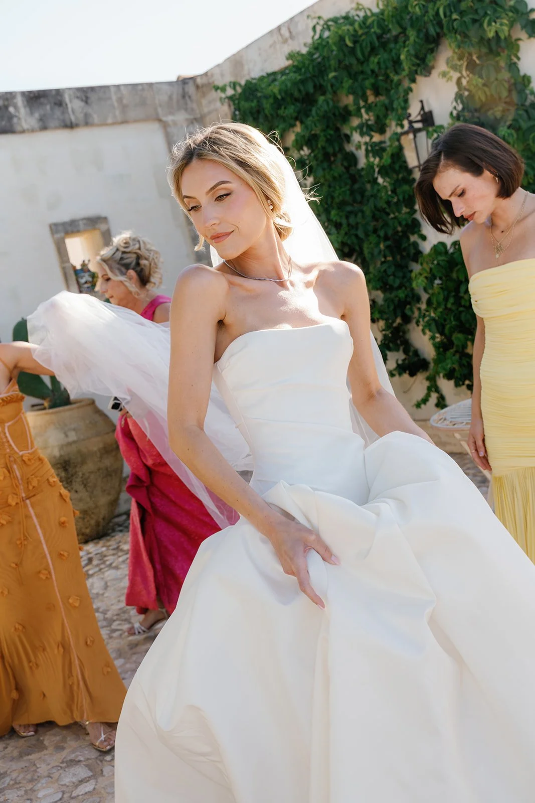 Bride in a white wedding gown standing outside with bridesmaids, some adjusting dresses and preparing for ceremony.