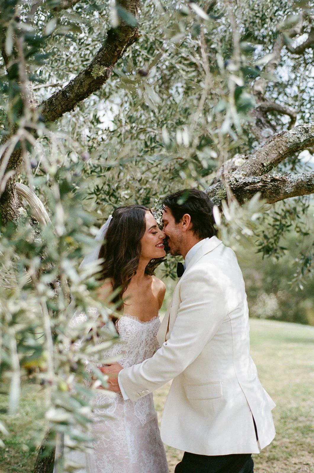 A bride and groom in wedding attire sharing a kiss under a tree with lush green leaves.