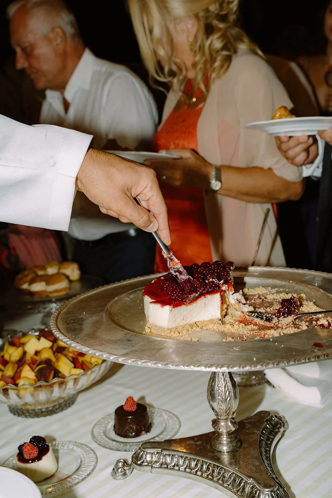 A person cutting a slice of cheesecake topped with red berries at a dessert table, with people holding plates in the background.