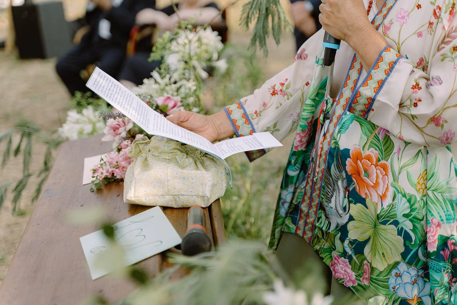A person wearing a colorful floral jacket holding a microphone and reading from a paper during a ceremony outdoors, with a table of flowers and notes in front.