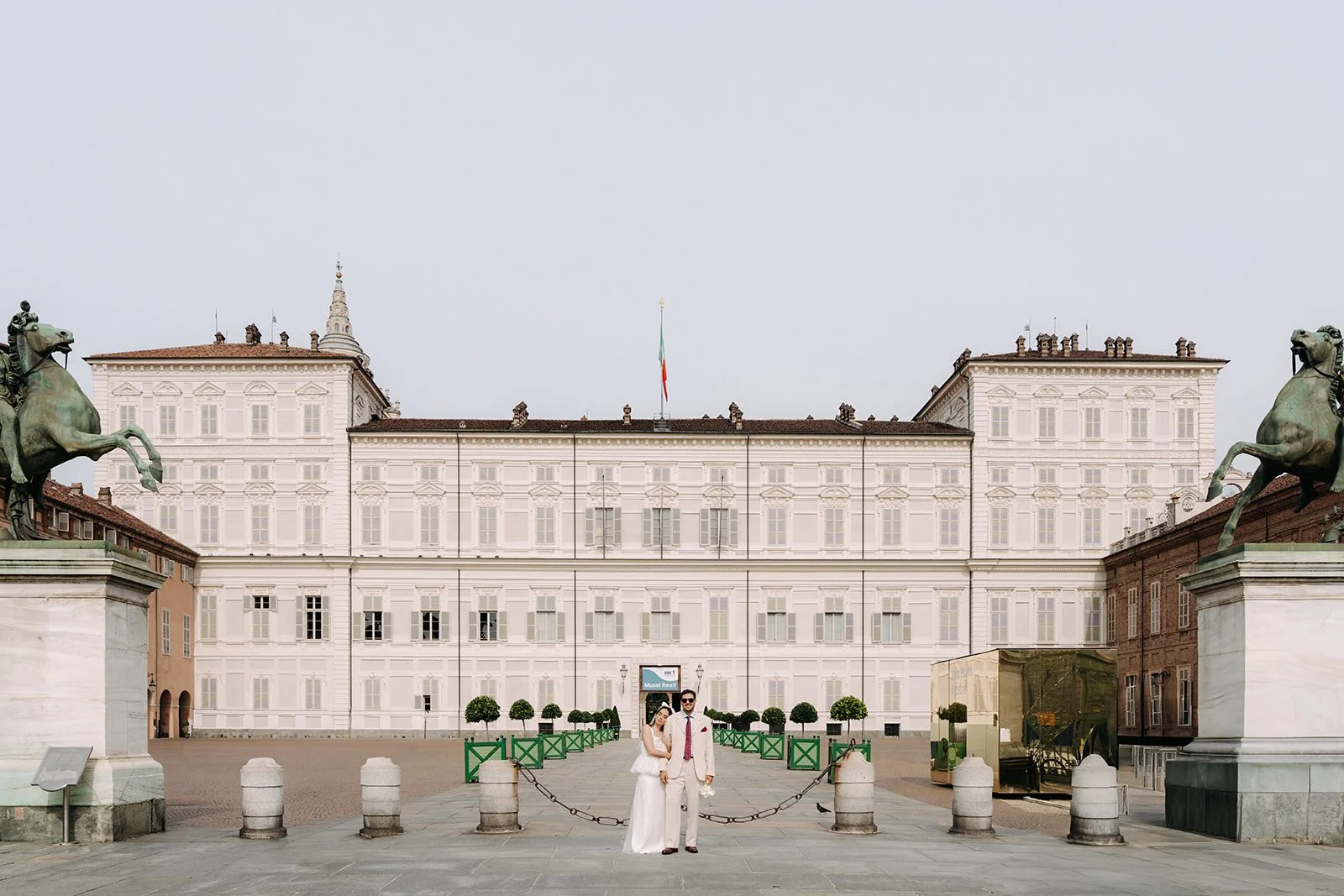 A couple dressed in formal white attire standing in front of a historic building with two large equestrian statues at each side.
