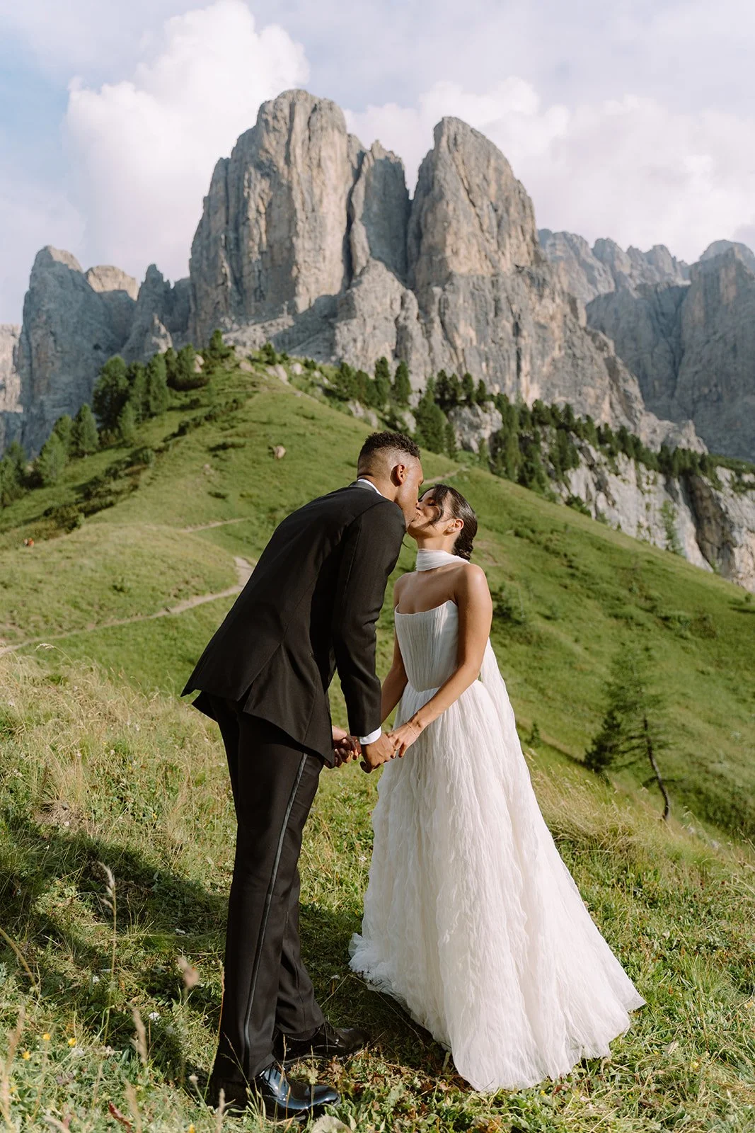 A newlywed couple in formal wedding attire sharing a kiss and holding hands outdoors on a grassy hillside with a mountain range in the background.