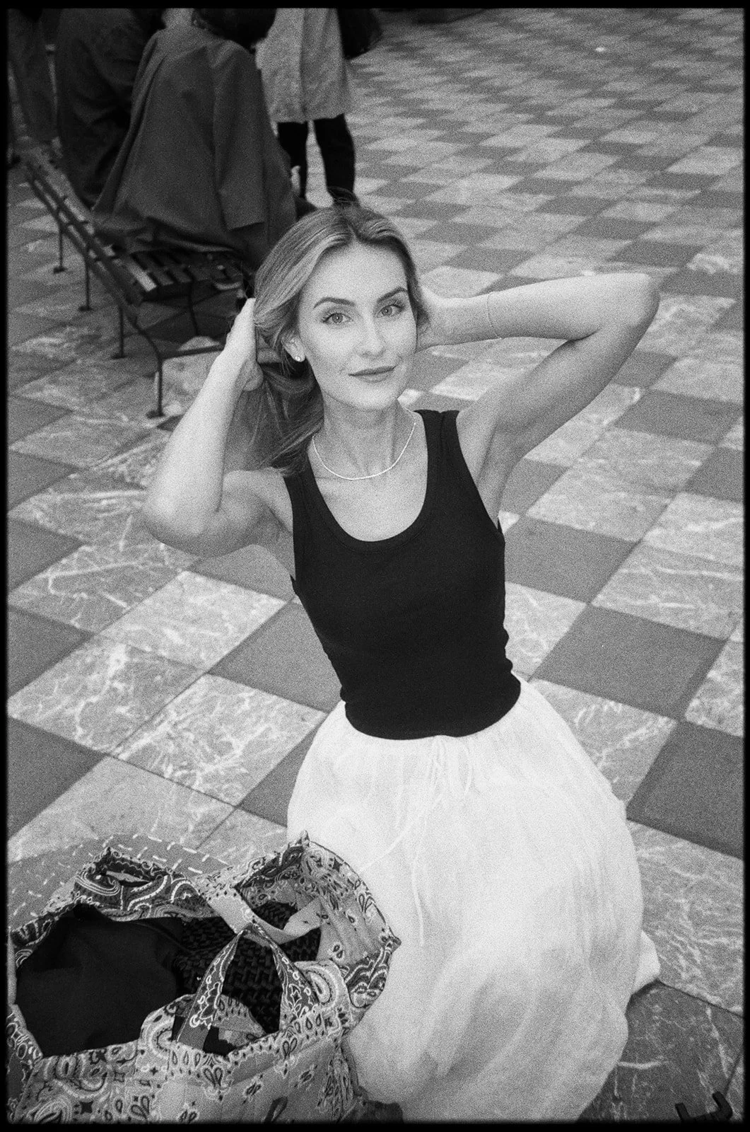 A woman sitting on the floor with her hands behind her head, smiling, wearing a black tank top and a light-colored skirt, next to a bag filled with various items