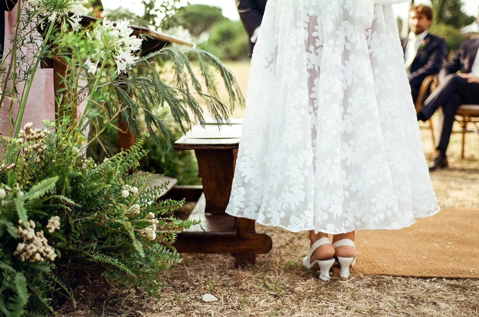 Close-up of a woman wearing a white lace dress and white high-heeled shoes, standing on a dirt ground with a brown mat, at an outdoor wedding ceremony. In the background, there are seated guests, some in formal suits, and greenery decorations.