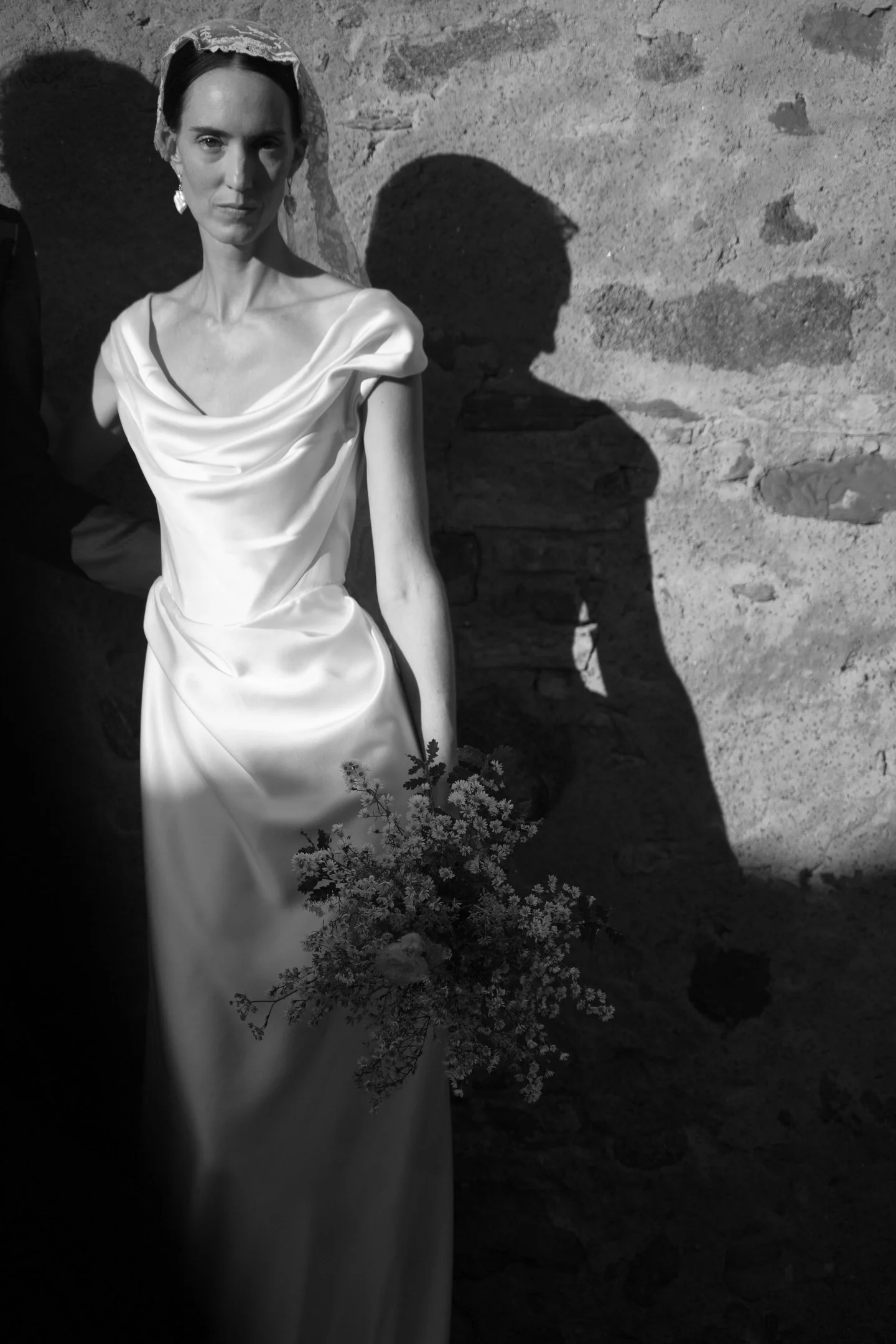 A woman in a long, elegant satin dress holding a bouquet of flowers standing against a stone wall. The sunlight casts a sharp shadow of her face and body on the wall.
