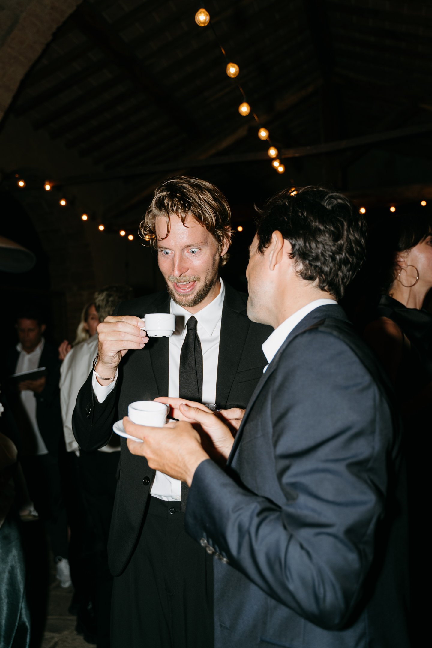 Two men dressed in formal suits and ties are engaging in a lively conversation, holding white teacups and saucers; one man has curly hair and a beard, displaying an animated facial expression, while the other has dark hair, with a bustling, dimly lit
