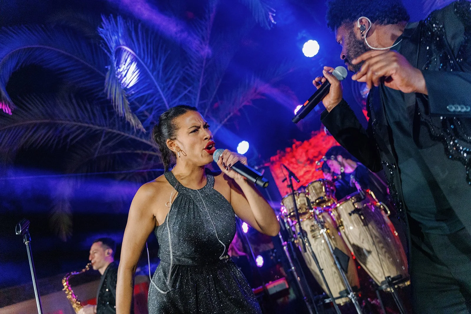 Two singers performing on stage with colorful lights and palm trees in the background. The woman on the left is wearing a sparkly black dress and holding a microphone, expressing emotion. The man on the right, wearing a black jacket, is also holding 