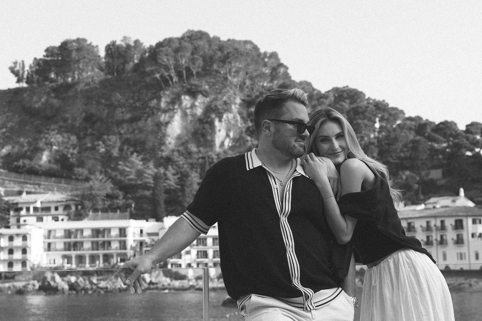 A black and white photo of a man and woman leaning on a railing by the water, with a hillside and buildings in the background.