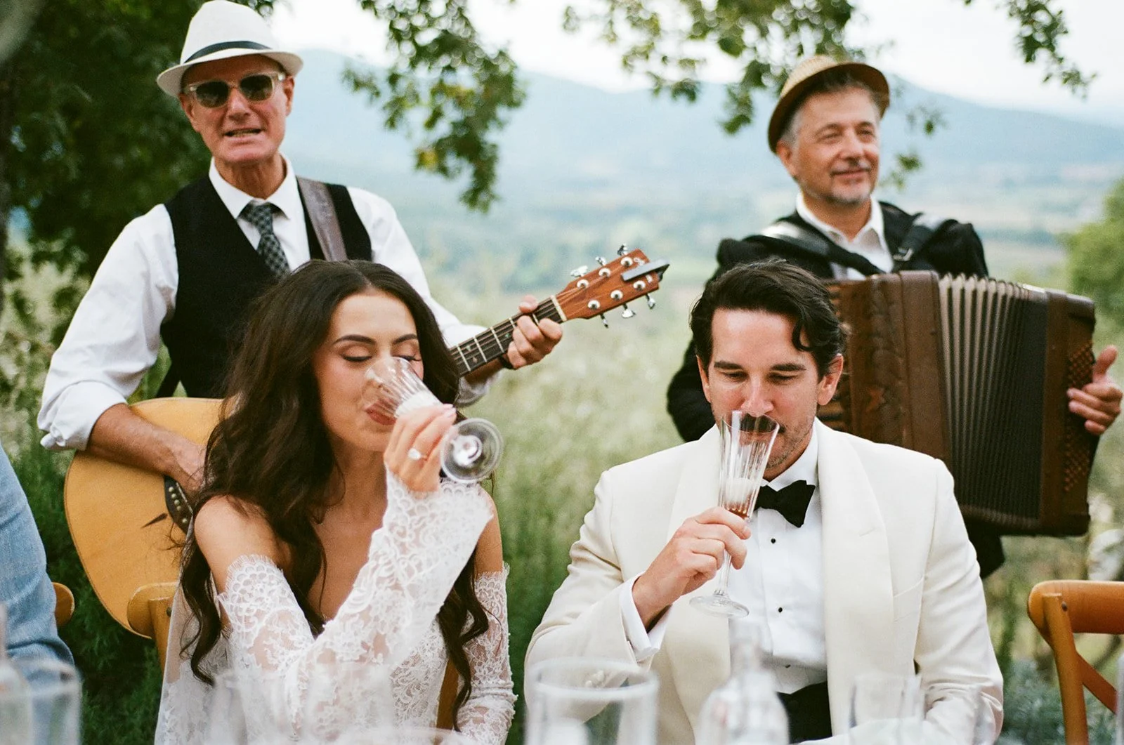 A bride and groom sitting at a table outdoors, drinking wine, while two men in vintage attire play musical instruments in the background, one with a guitar and the other with an accordion.