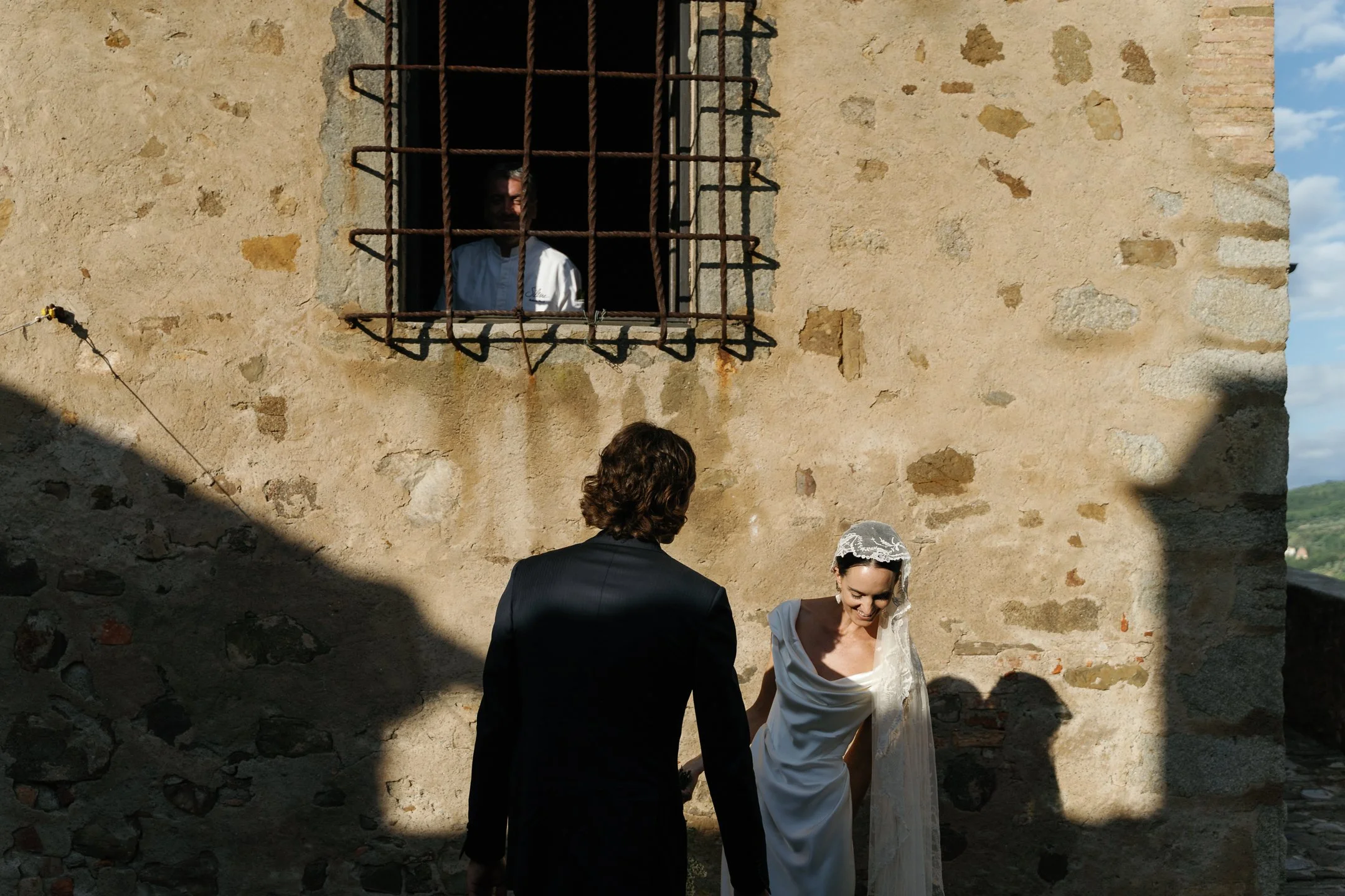 A bride and groom holding hands outside a rustic stone building with a barred window, with a woman observing from the window, in a setting with natural sunlight and shadows.