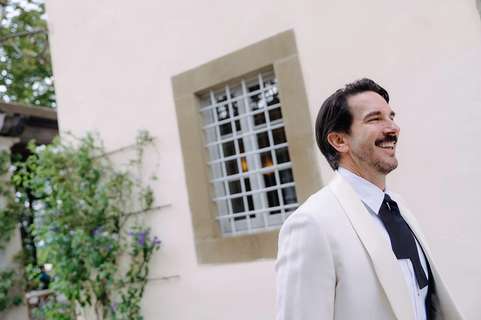 A man dressed in a white tuxedo with a black tie smiling outdoors near a building with a window and greenery in the background.