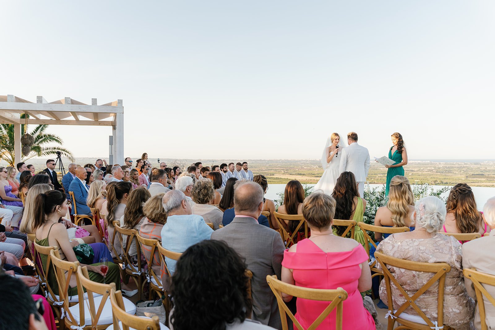 Outdoor wedding ceremony with bride and groom standing under a white wooden arch, officiant and bridesmaid reading, guests seated facing the couple, scenic landscape in the background, clear sky, tropical palm trees on the left.