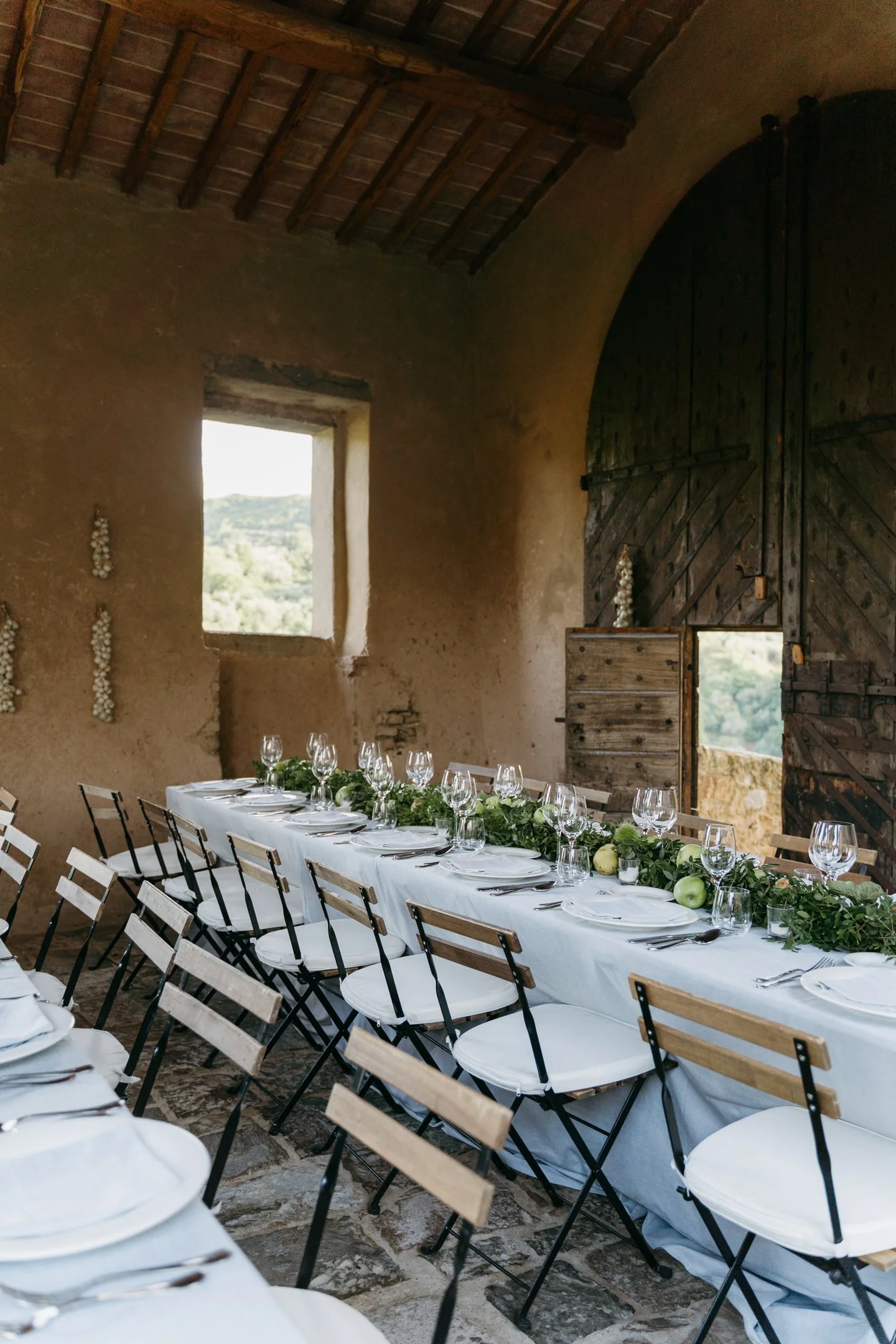 An elegantly set dining table with white tablecloth, glasses, plates, and cutlery, decorated with green foliage and white flowers, in a rustic stone-walled room with large wooden doors and a window showing a scenic outdoor landscape.