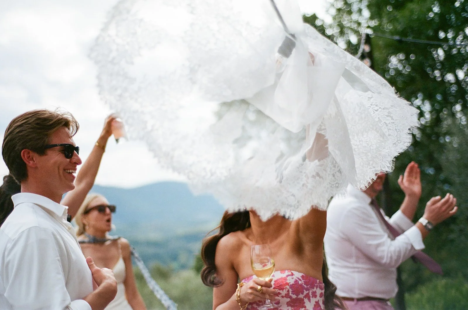 People celebrating outdoors, one woman with a lace umbrella and another man smiling, holding a drink.