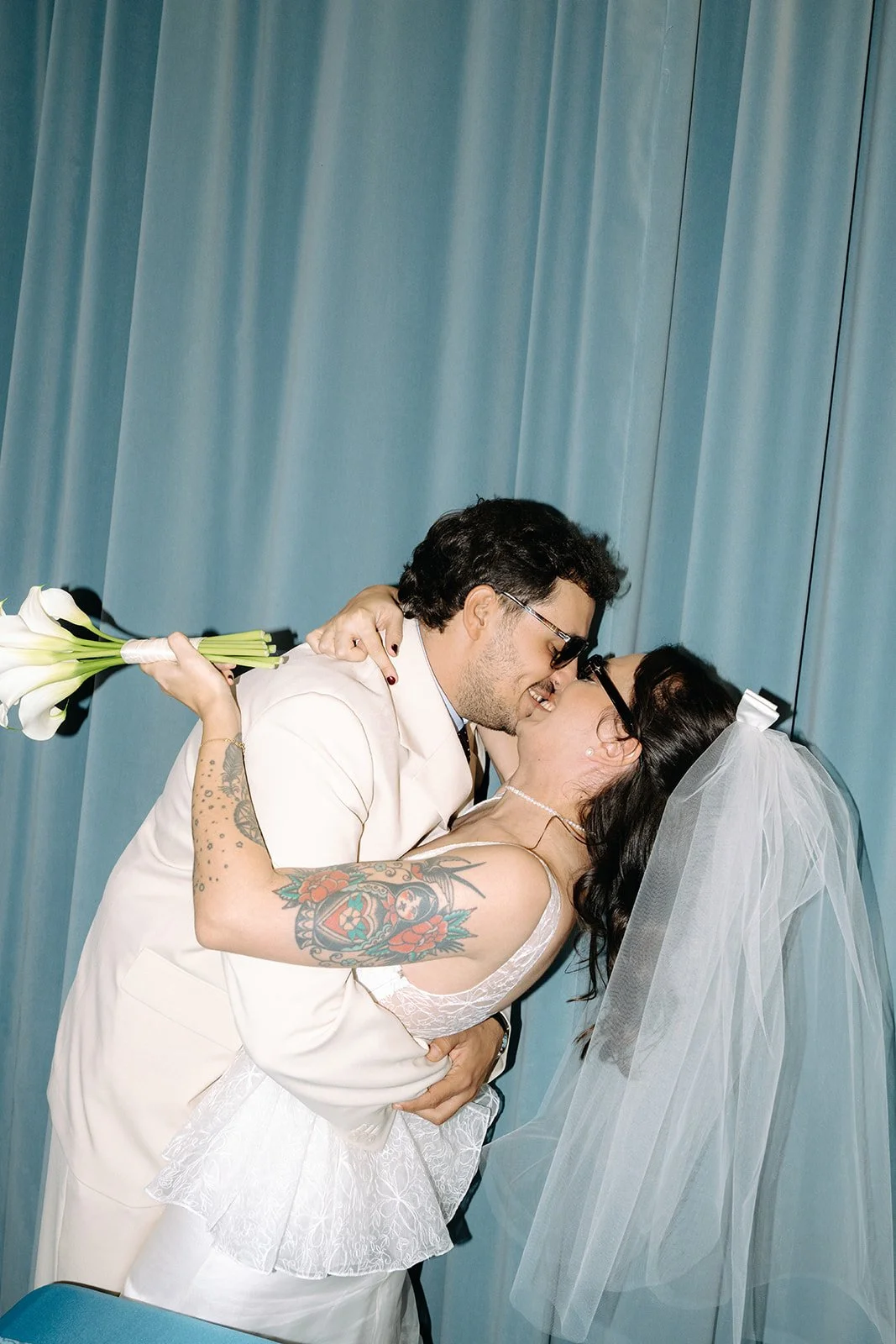 A bride and groom share a kiss at their wedding, with the groom holding a bouquet of white calla lilies and both wearing glasses. Blue curtain backdrop.