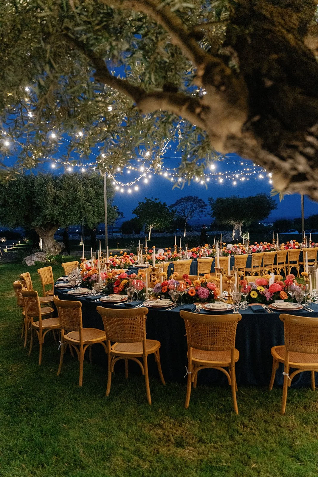 An outdoor dinner setup for a celebration at night, featuring a long table with pink and orange floral centerpieces, candles, and place settings. String lights hang overhead, and trees are visible in the background.