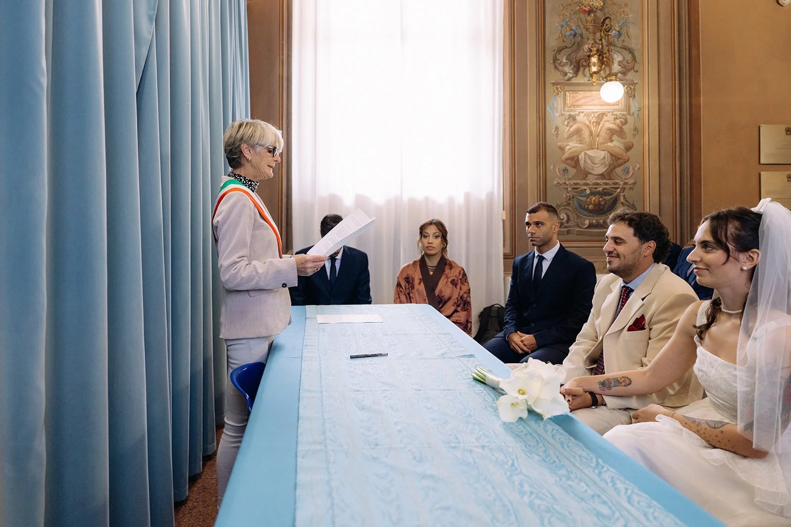 A wedding ceremony taking place in an ornate room with a woman officiating, three seated guests, and a bride holding a bouquet of white flowers, all sitting at a light blue table.