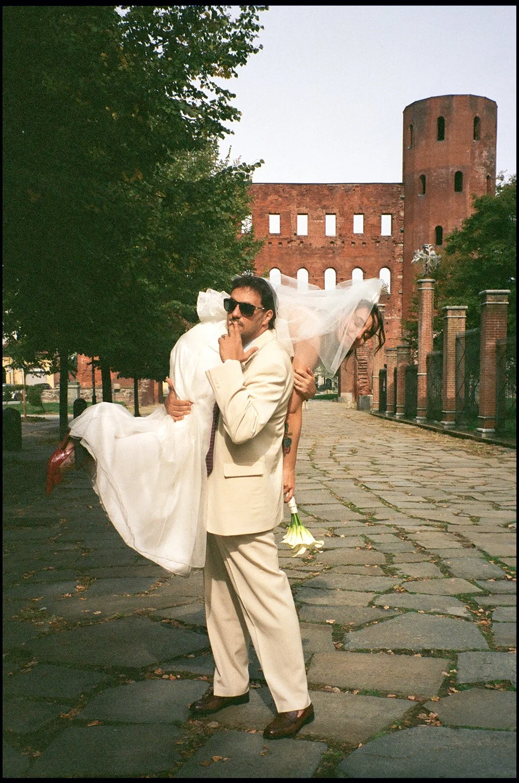 A man in a beige suit holding a woman in a wedding dress and veil, with the woman holding a bouquet of white flowers, stands on a cobblestone street with an ancient brick building and tower in the background.
