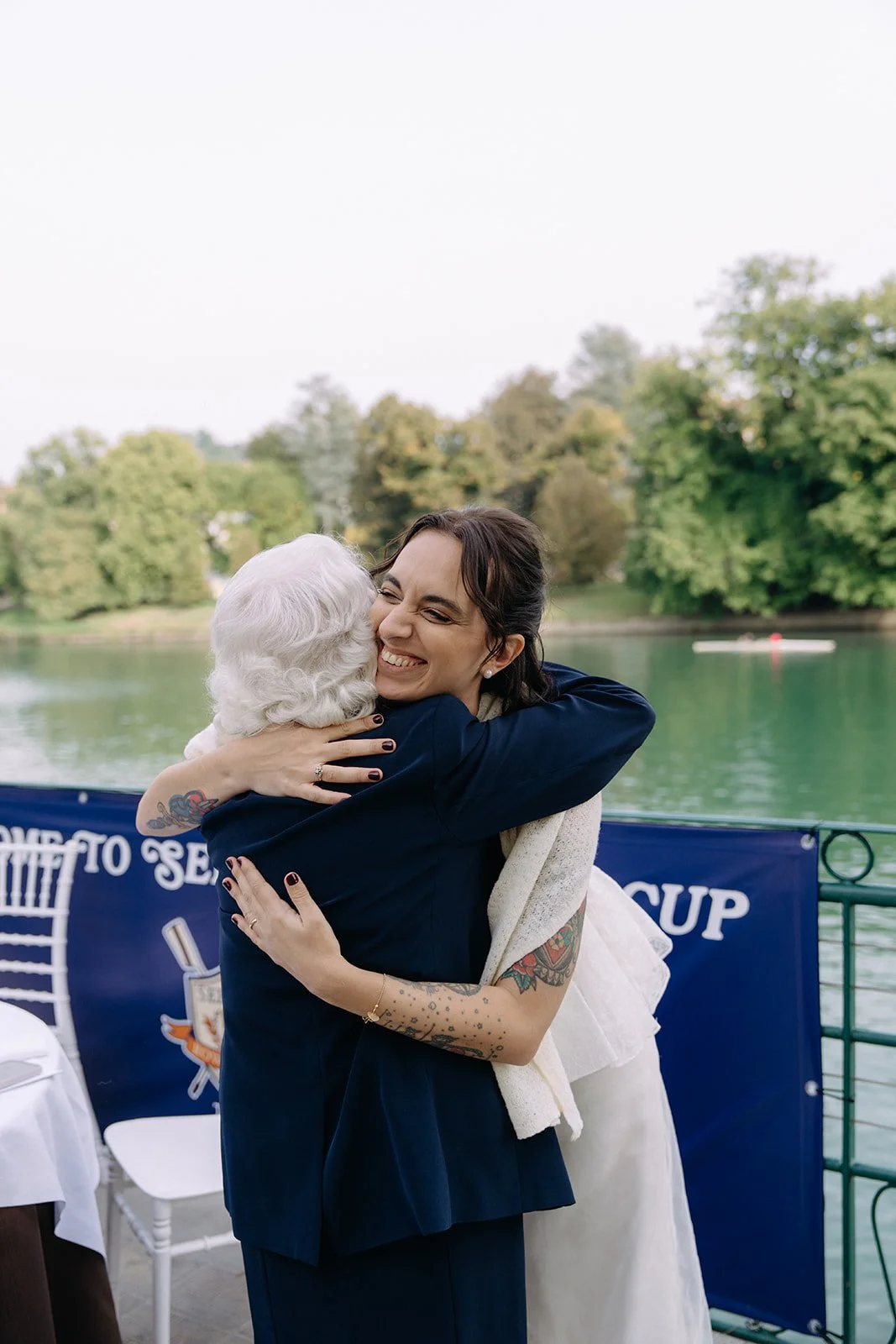 Two women hugging and smiling outdoors near a river, with trees in the background.