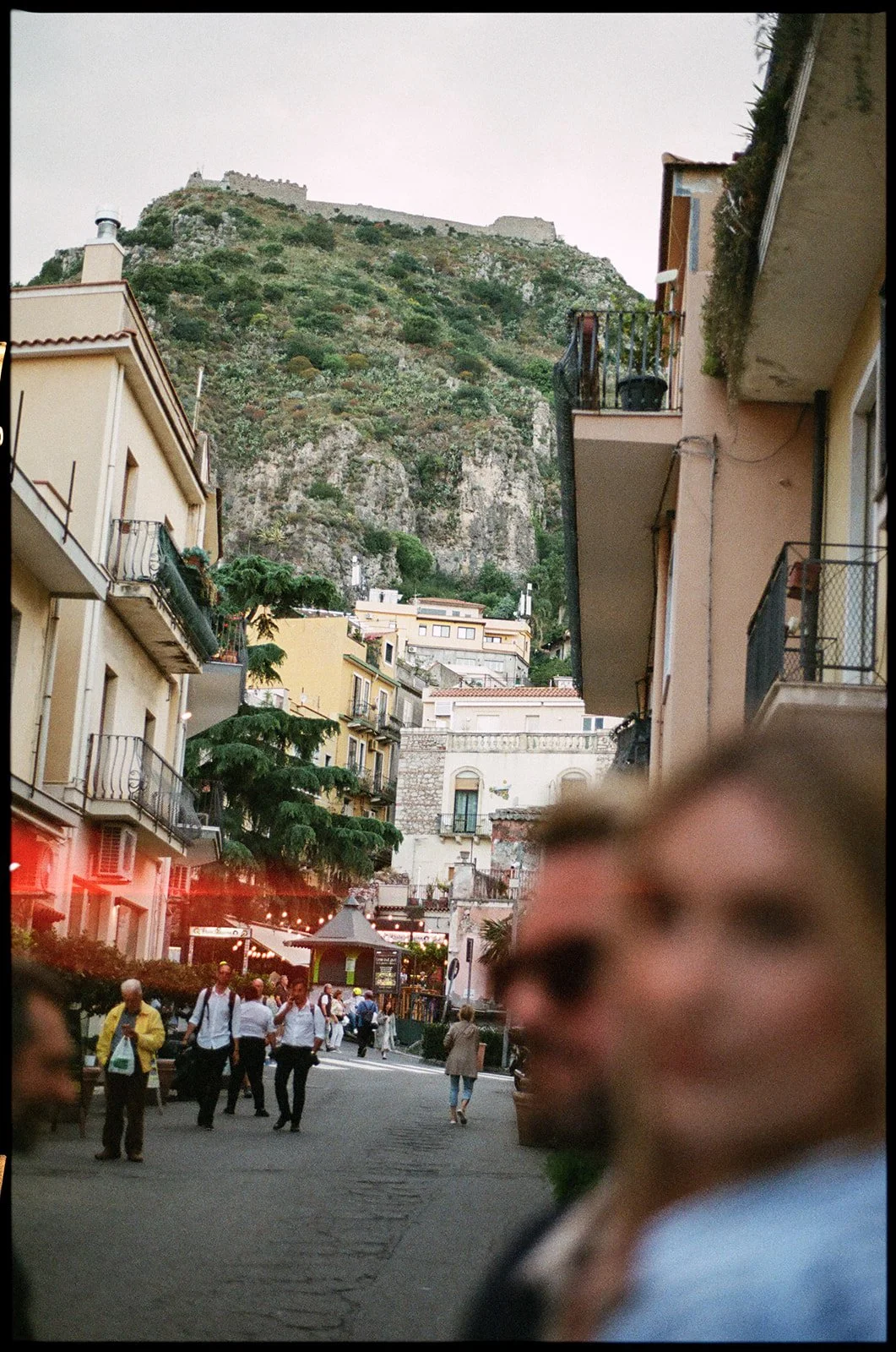 Blurred person in foreground with a street scene and colorful buildings, hillside with castle above in the background.