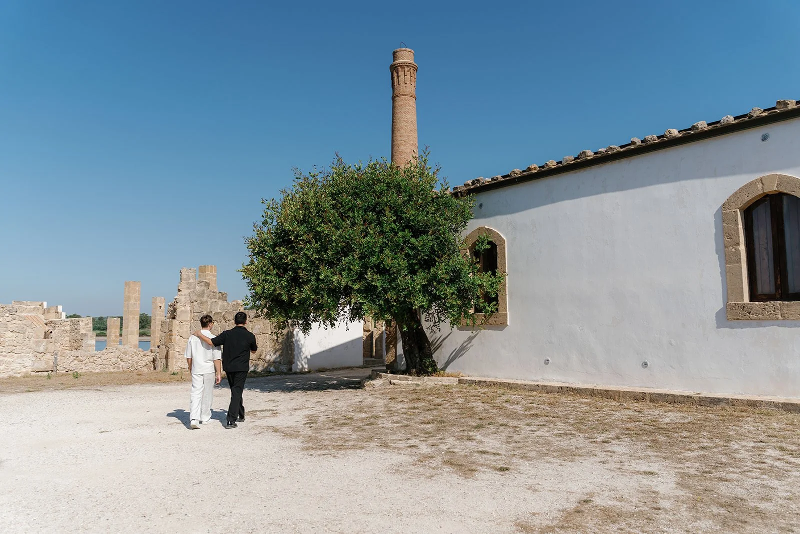 Two people walking arm in arm on a dirt path near a white building with arched windows, a large green tree, and a tall brick chimney under a clear blue sky.