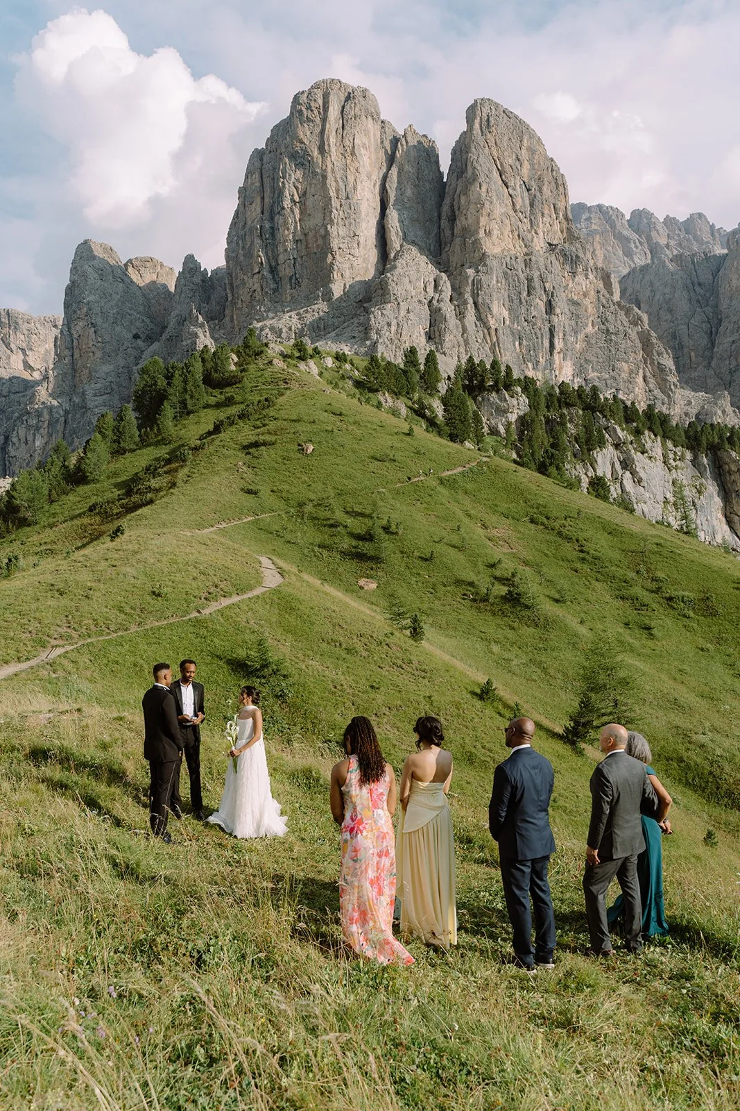 A wedding ceremony taking place outdoors on a grassy hillside with a mountain range in the background. The bride is in a white wedding dress, and the groom and officiant are in suits, with guests standing nearby.