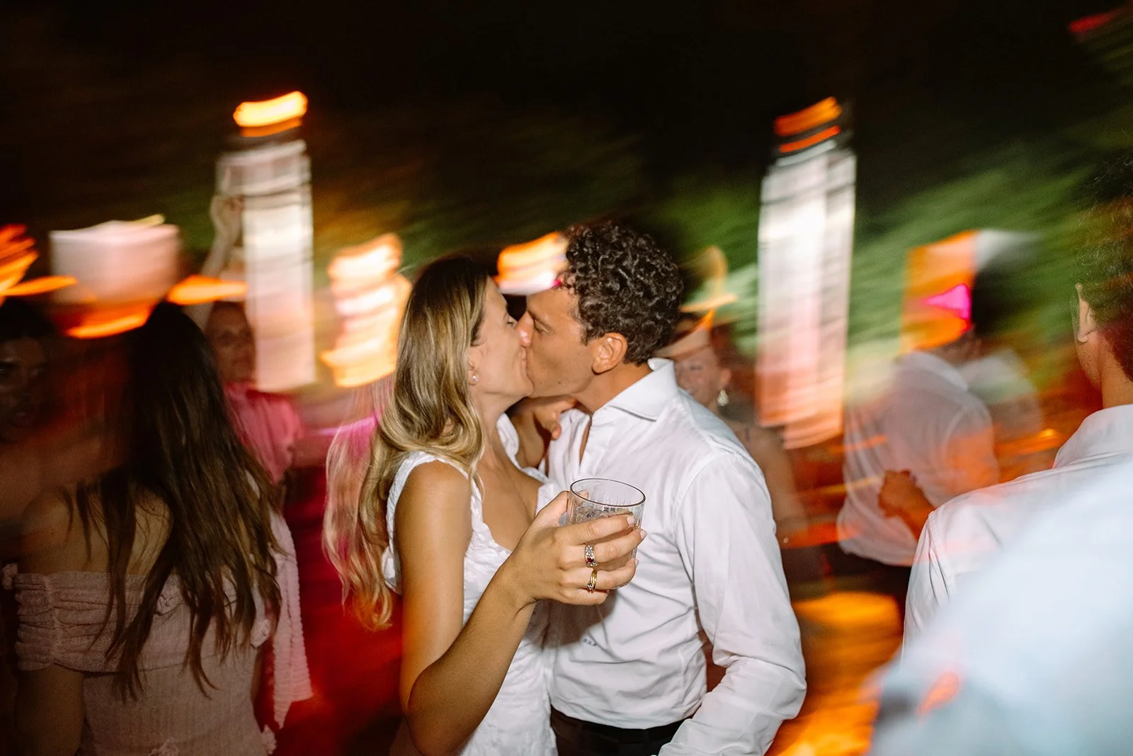 Couple kissing at party with colorful lights and blurred guests in background.