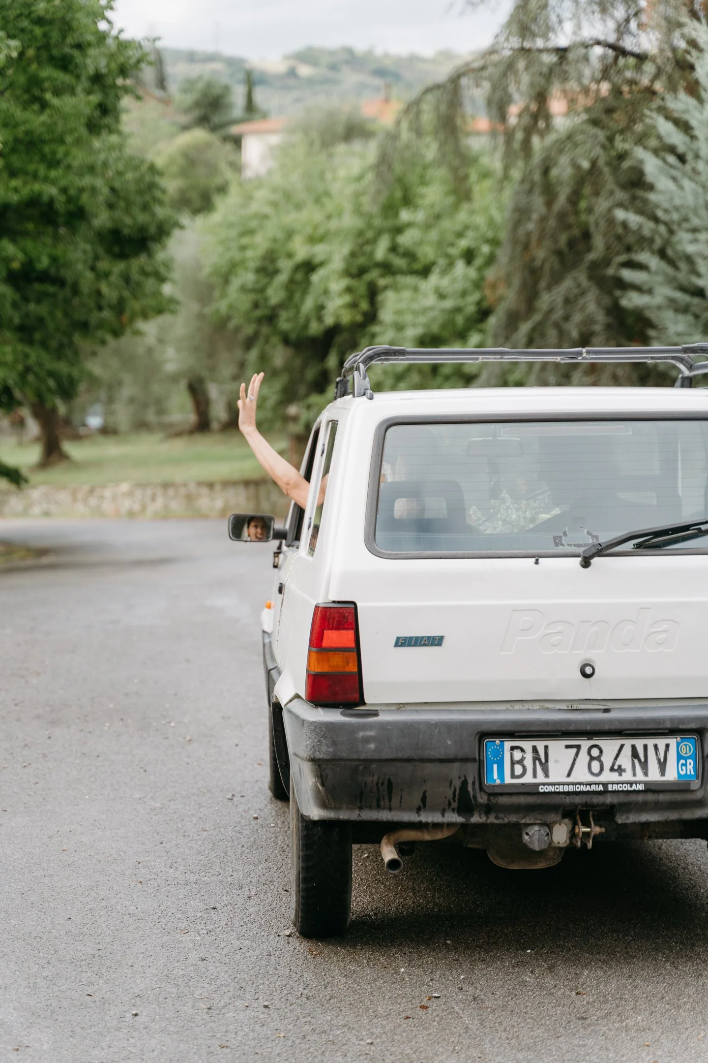 Rear view of a white Fiat Panda with a person's arm and hand waving out of the passenger window, parked on a paved road in a green, rural area.