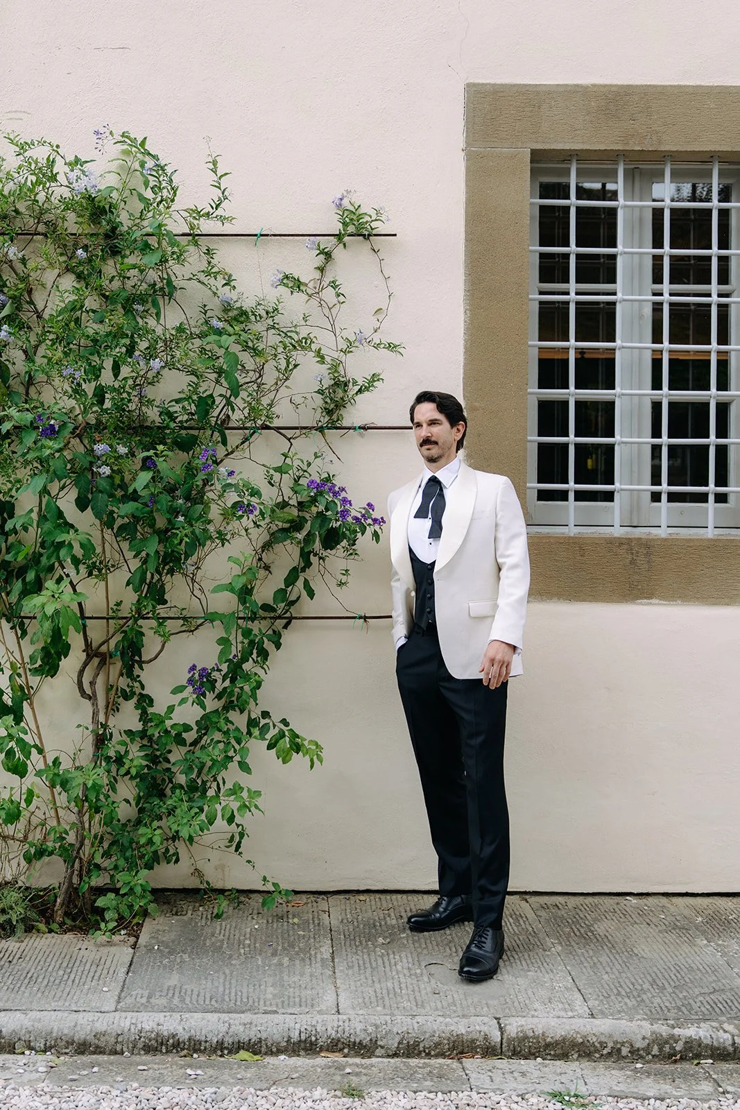 A man in formal attire standing outdoors next to a wall with a window and green plants with purple flowers.