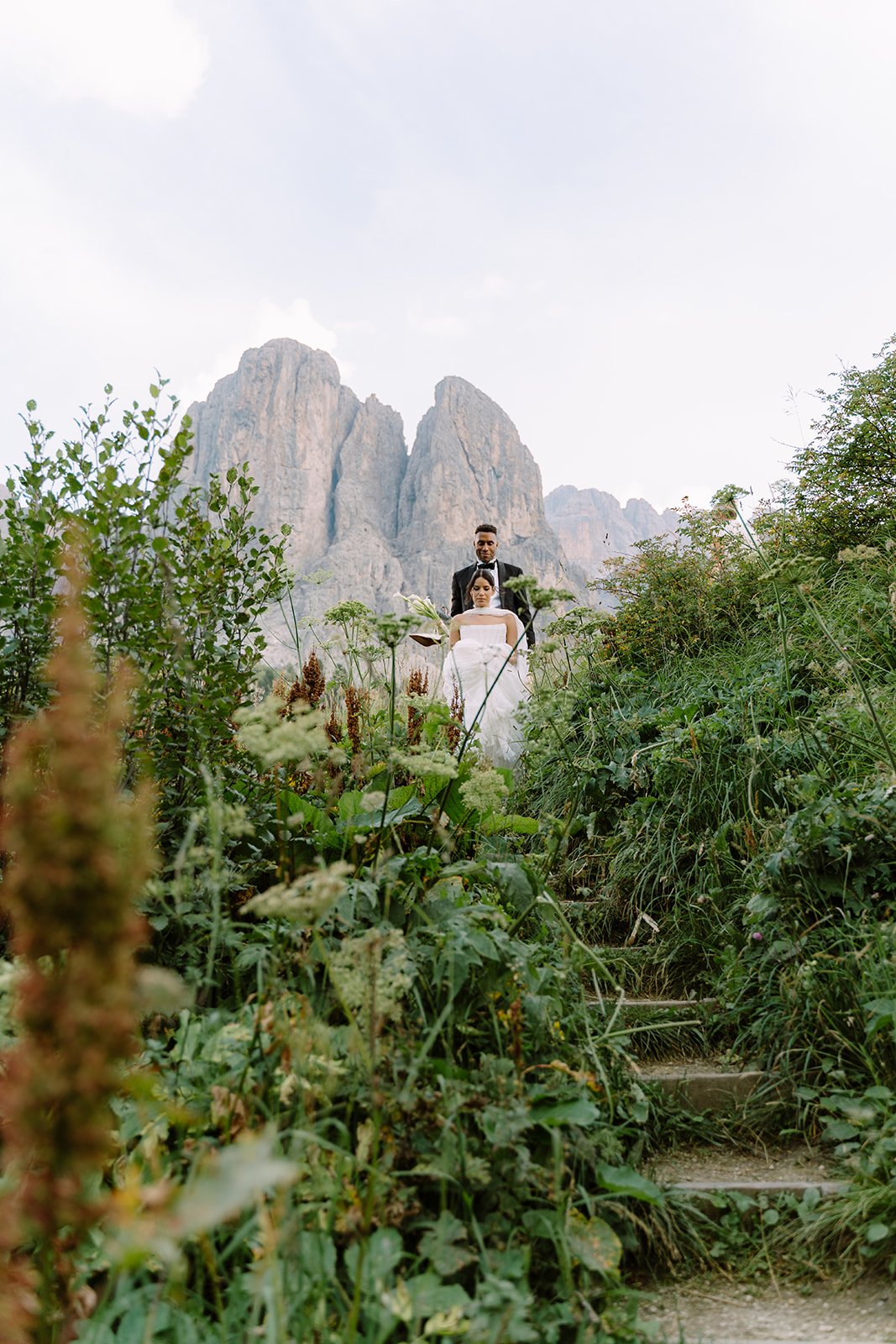 A bride and groom in wedding attire standing on natural stone stairs in a lush, green outdoor setting with mountains in the background.