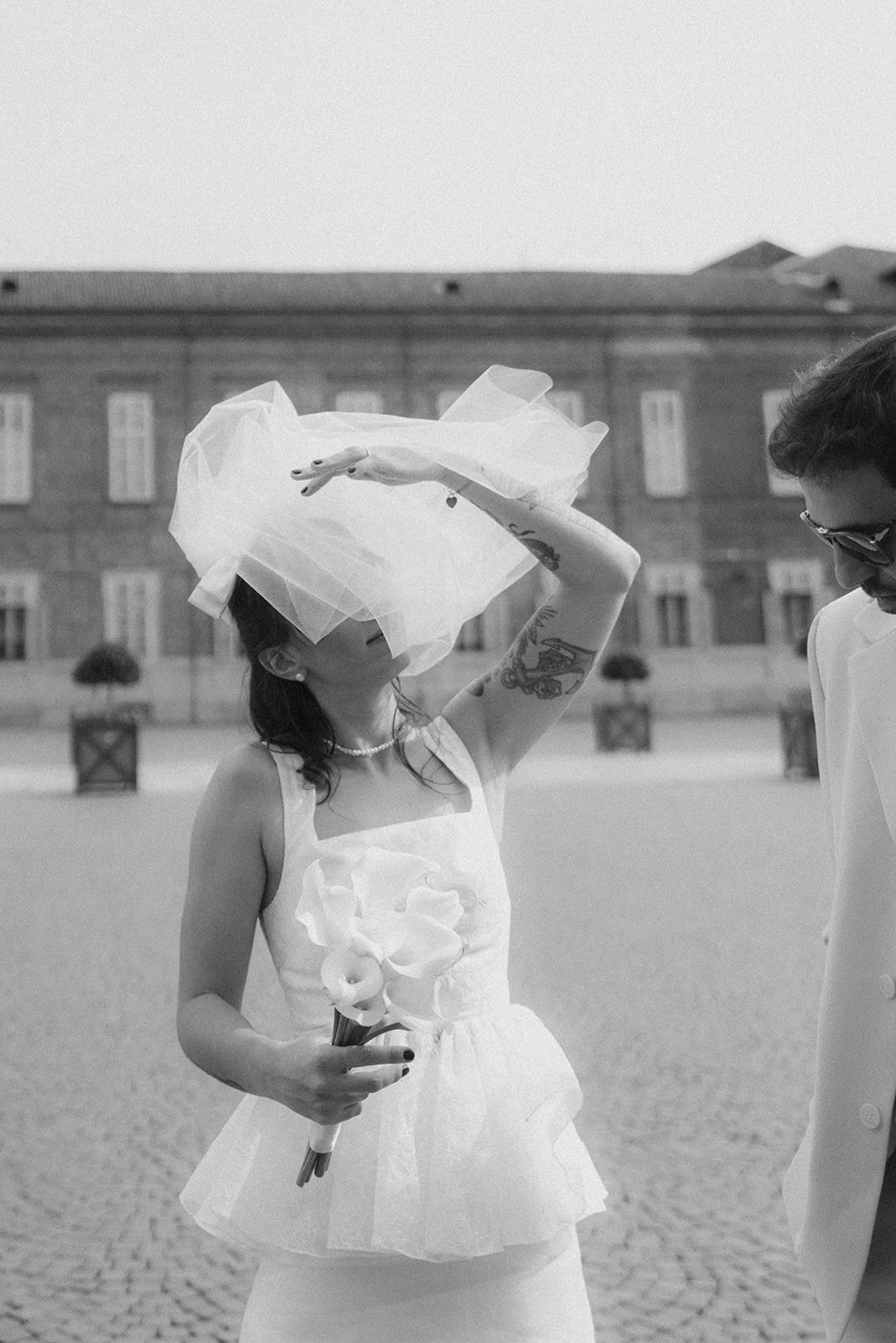 A woman wearing a white dress and a necklace, holding a bouquet, shielding her face with a tulle veil during an outdoor event, with a building and cobblestone ground in the background.