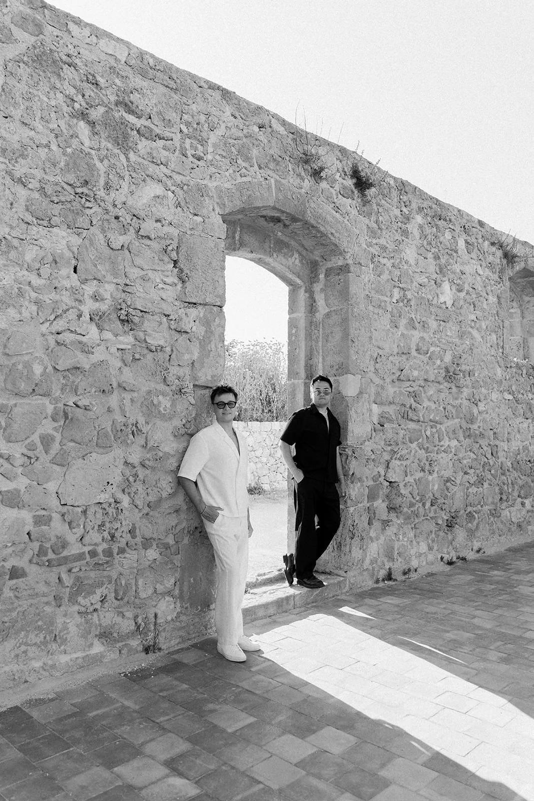 Two men standing in a doorway of an old stone wall, one in a white outfit and the other in a dark outfit, outside on a sunny day.