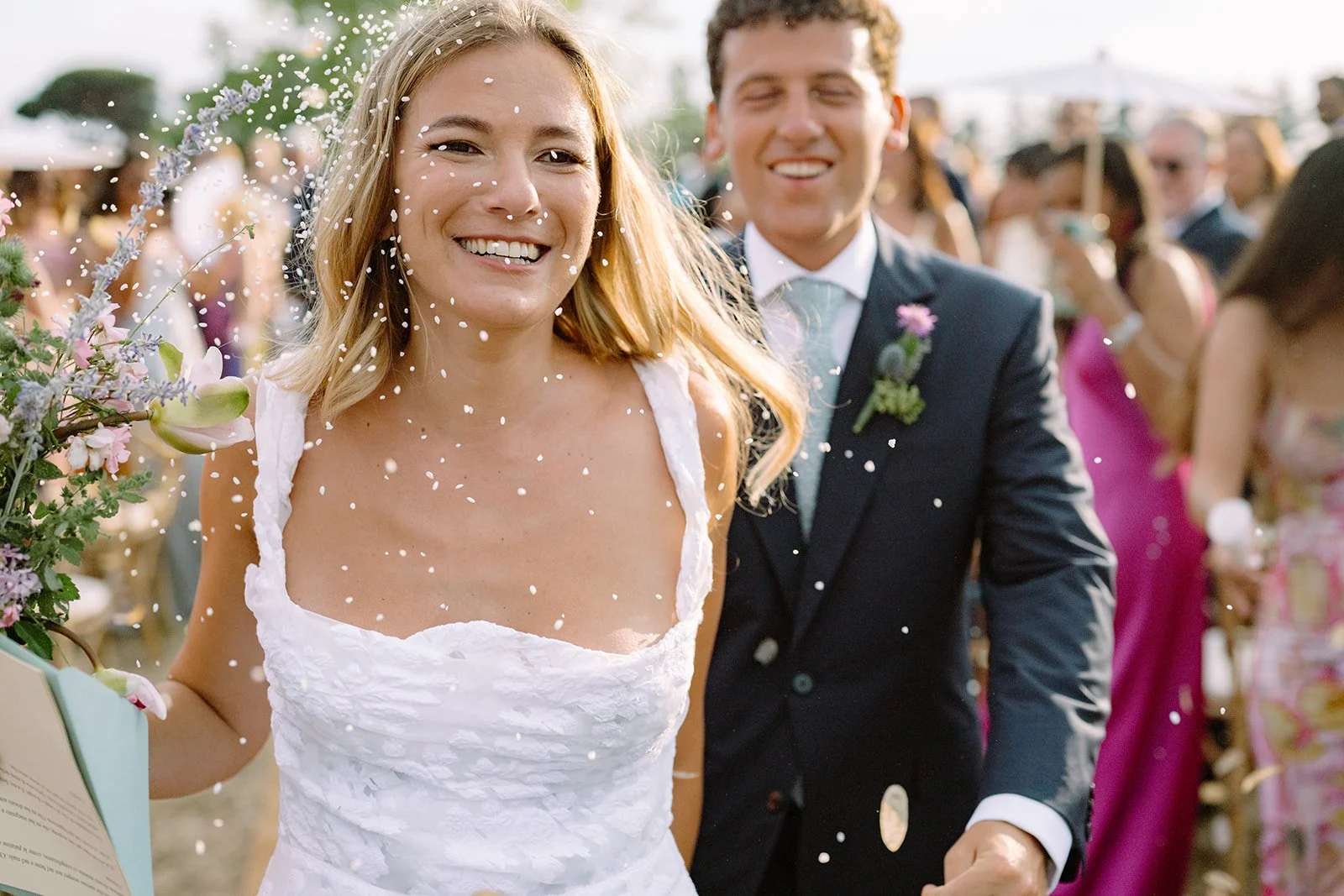A smiling woman in a wedding dress holding a bouquet, with confetti falling around her at an outdoor wedding celebration, while a man in a suit walks behind her surrounded by guests.