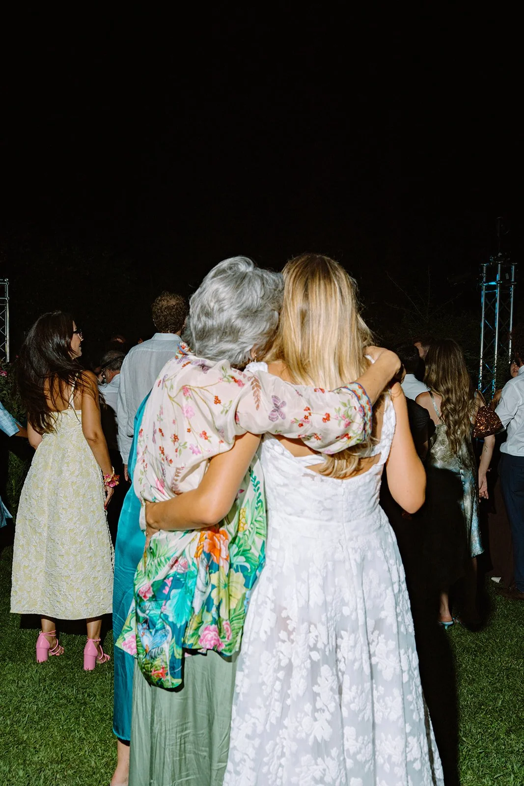 People hugging at a night outdoor event, with others mingling in the background.