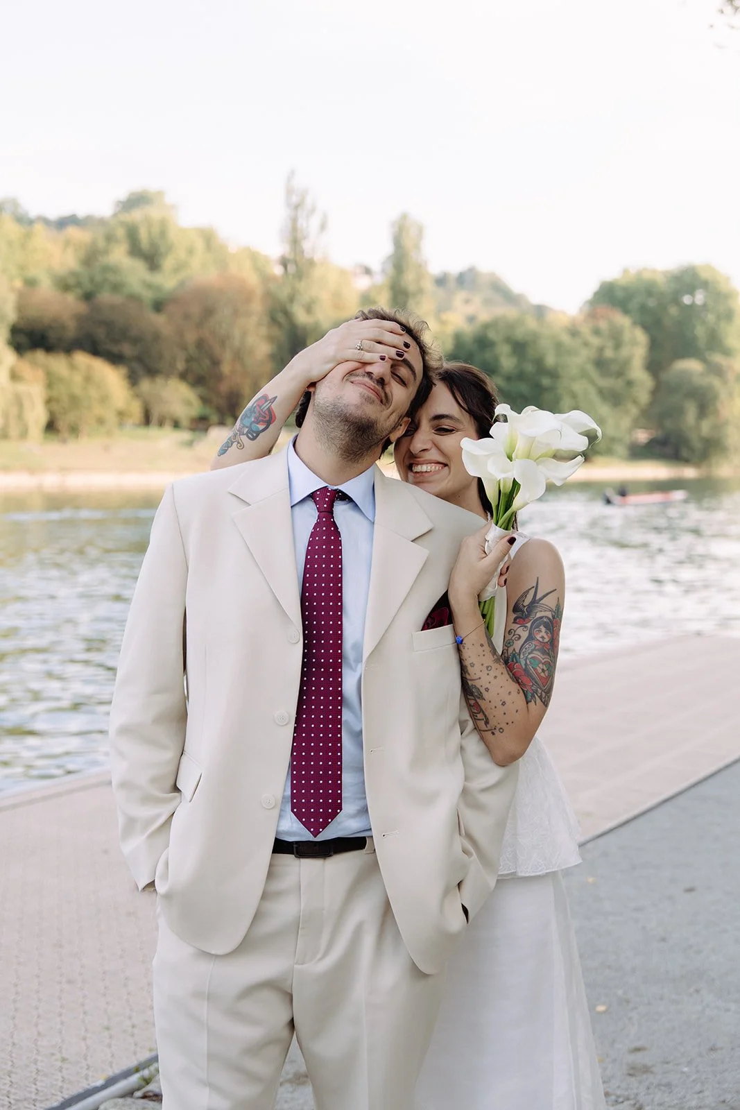 A happy couple near a river, with the woman holding white lilies and touching the man’s face, both smiling and enjoying a joyful moment.