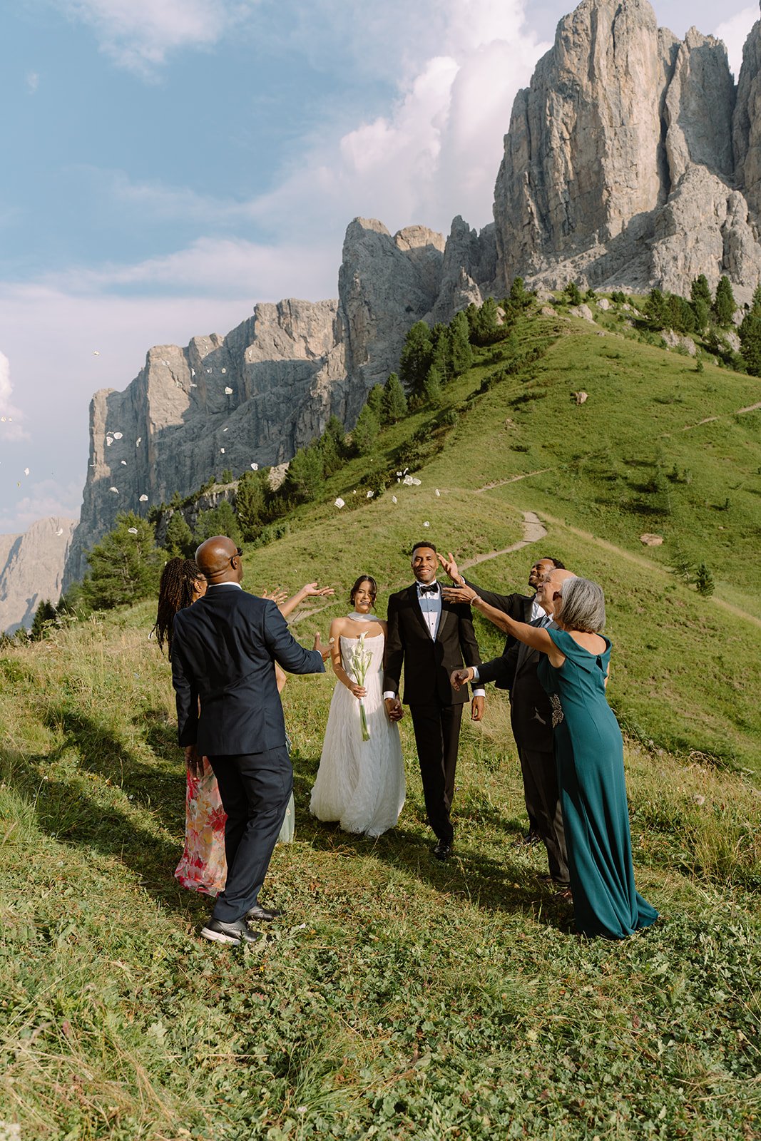 A wedding ceremony taking place outdoors in a lush green mountain landscape with a rocky mountain in the background. The bride, groom, and several guests are engaging joyfully, with some throwing flower petals.