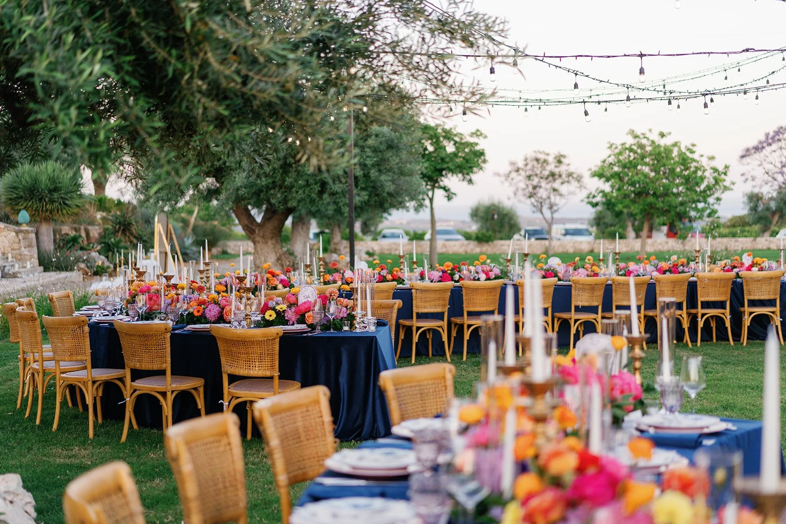 Outdoor wedding reception setup with long tables covered in navy blue tablecloths and decorated with colorful floral centerpieces, tall white candles, and elegant glassware, surrounded by wooden chairs on a grassy area with trees and string lights ov