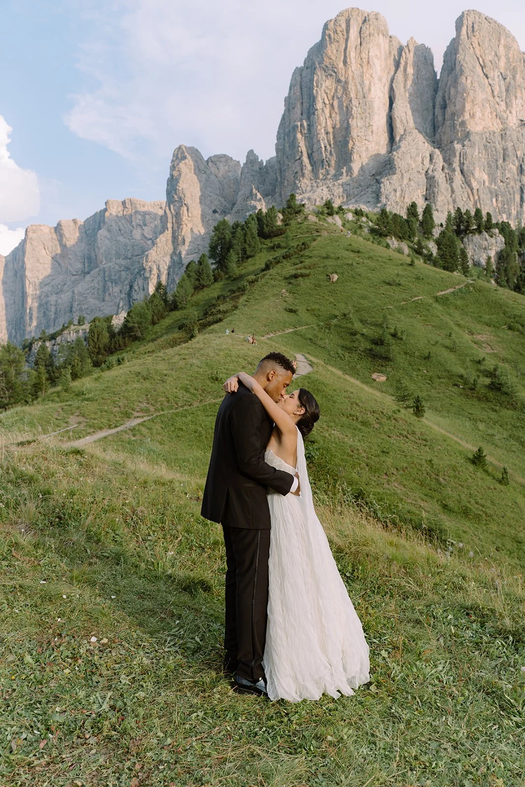 A bride and groom embracing in an outdoor mountain setting, with green hills and rocky cliffs in the background.