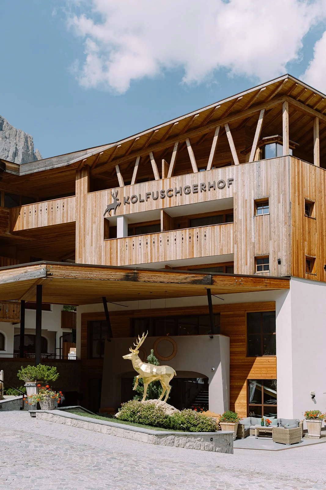 Wooden hotel with the name 'KOLFUSCHERHOF' and a deer sculpture in front, outdoor seating area with lounge chairs, and mountains in the background under a partly cloudy sky.