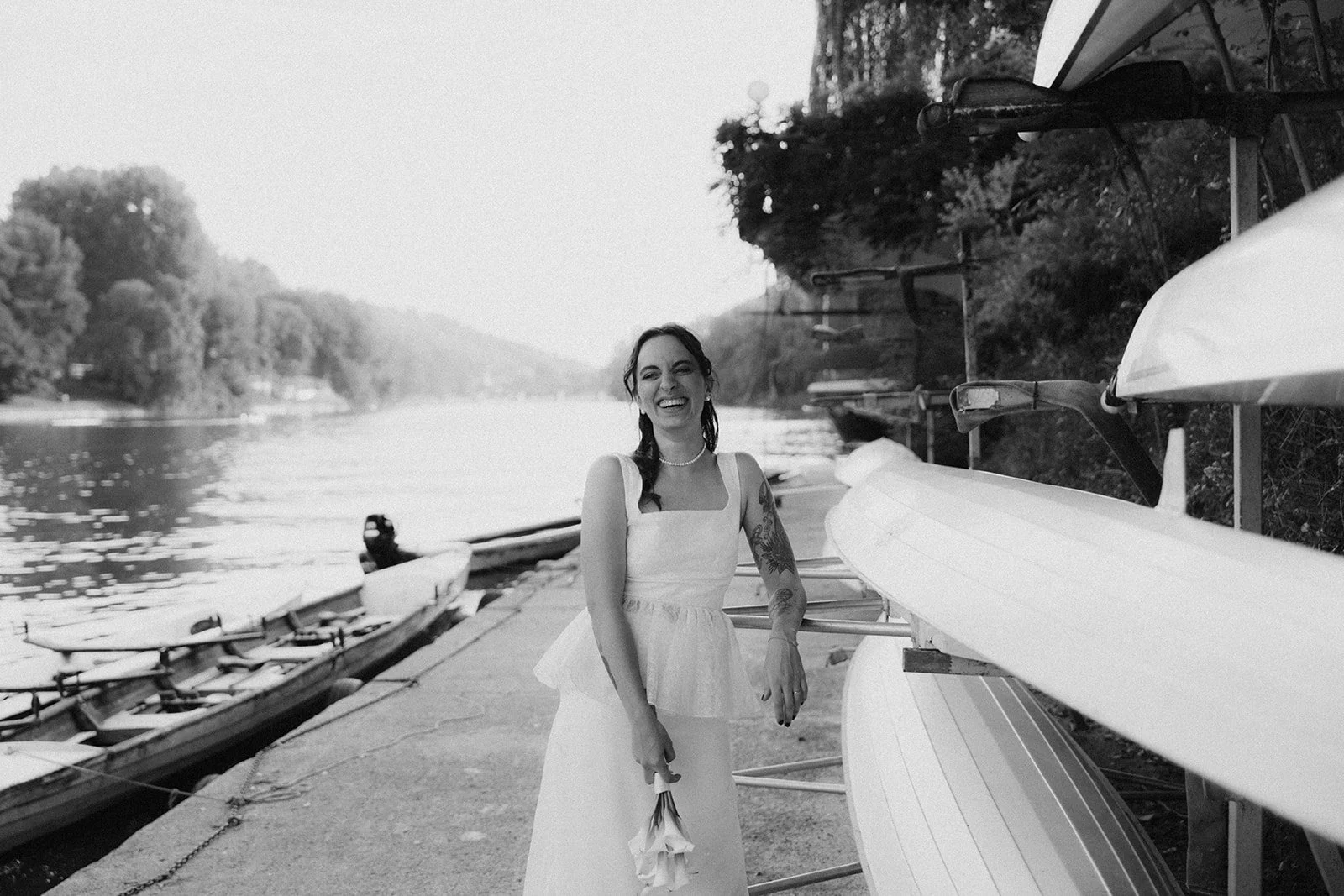 A woman in a white dress holding a bouquet of flowers standing beside kayaks near a river on a dock with trees in the background.