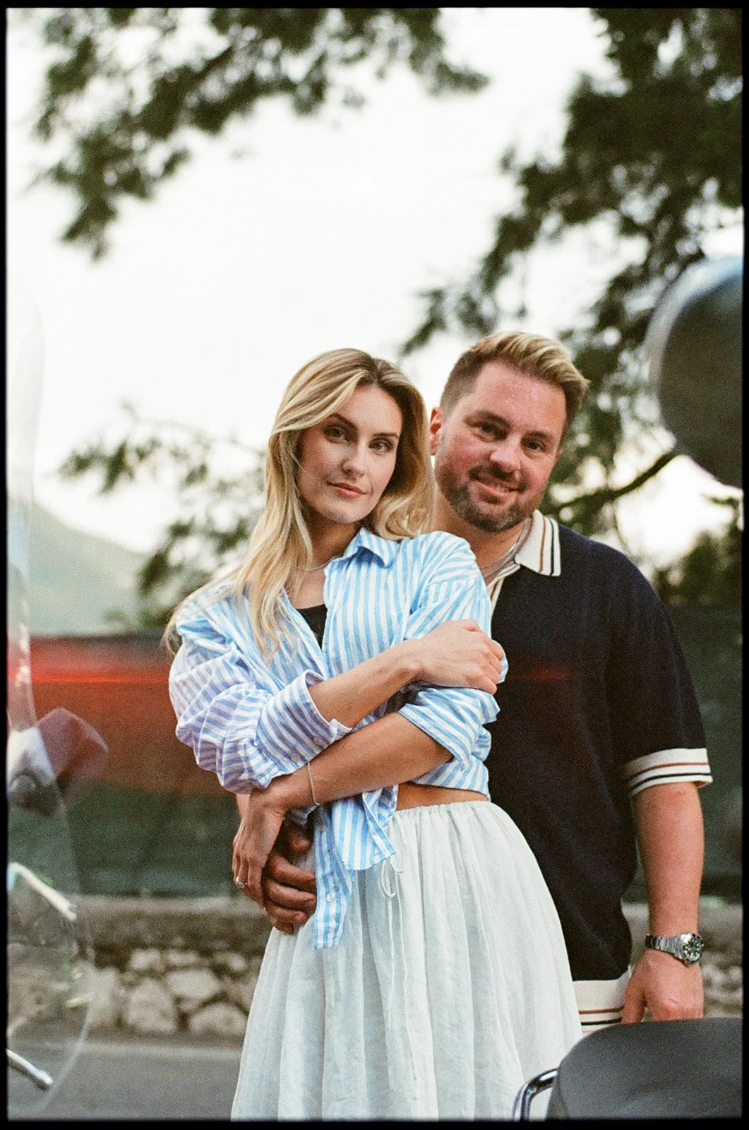 A young woman with blonde hair in a blue and white striped shirt standing in front of a young man with light brown hair in a black shirt. They are outdoors with trees in the background.