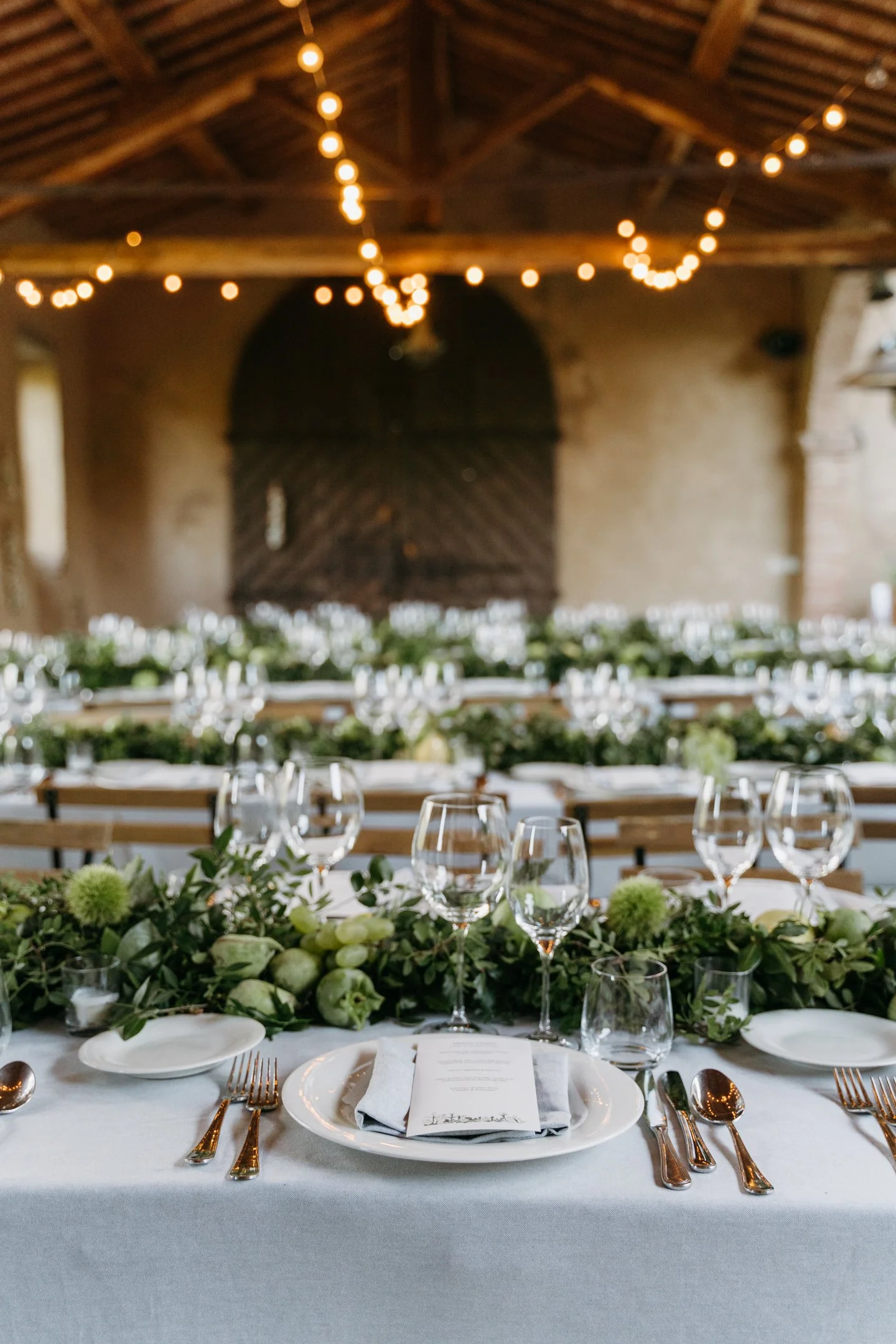 Elegant wedding reception table with white tablecloth, greenery, white flowers, wine glasses, and silverware, set in a rustic venue with string lights hanging from the wooden ceiling.