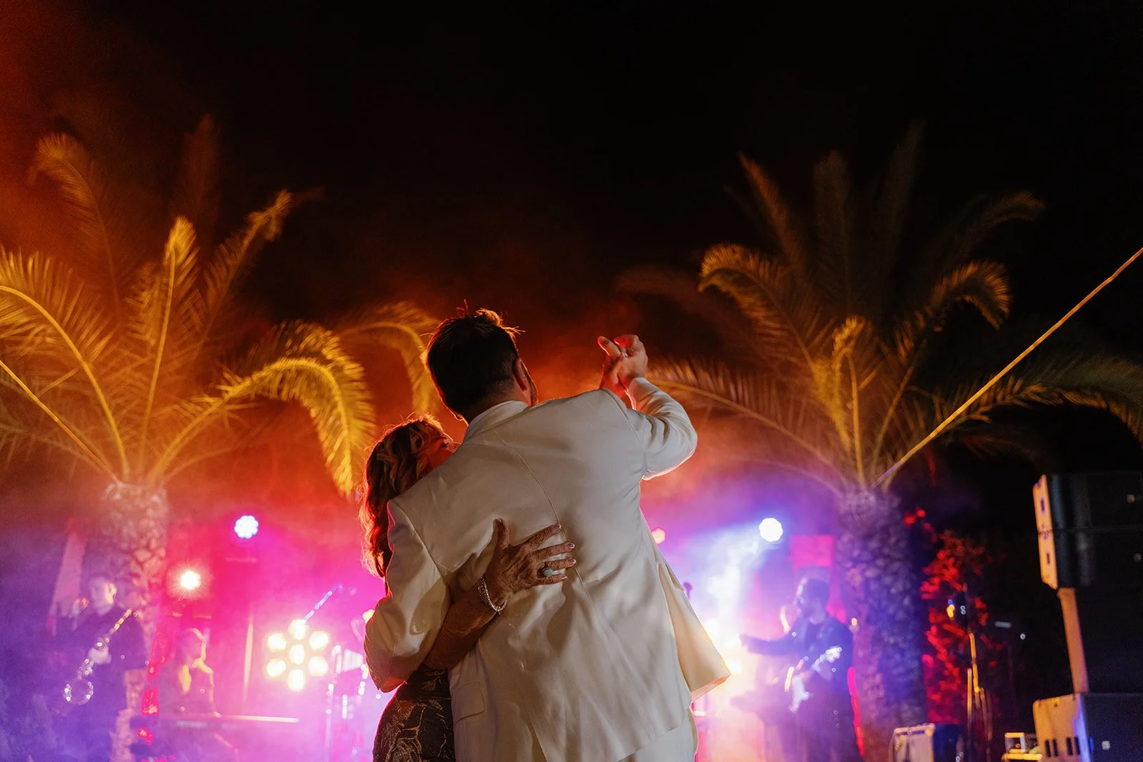 A couple dancing at an outdoor event at night, with palm trees and colorful stage lights in the background.