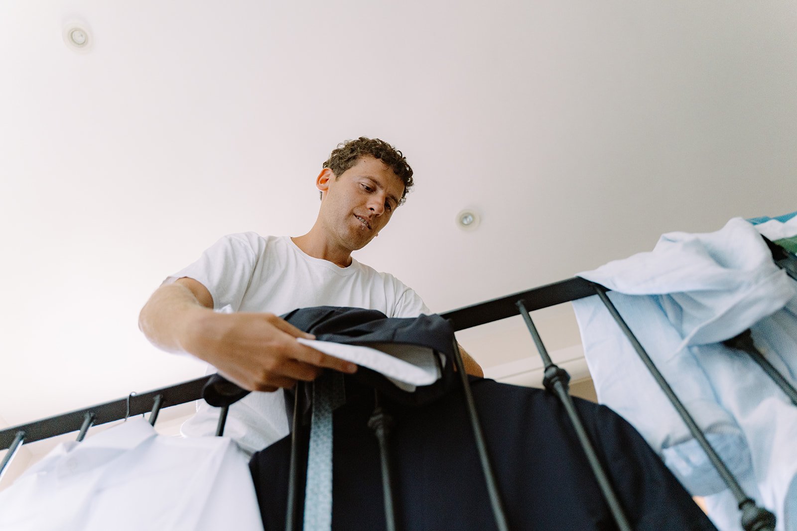 Man wearing a white shirt hanging clothes on a bed with a black metal frame.