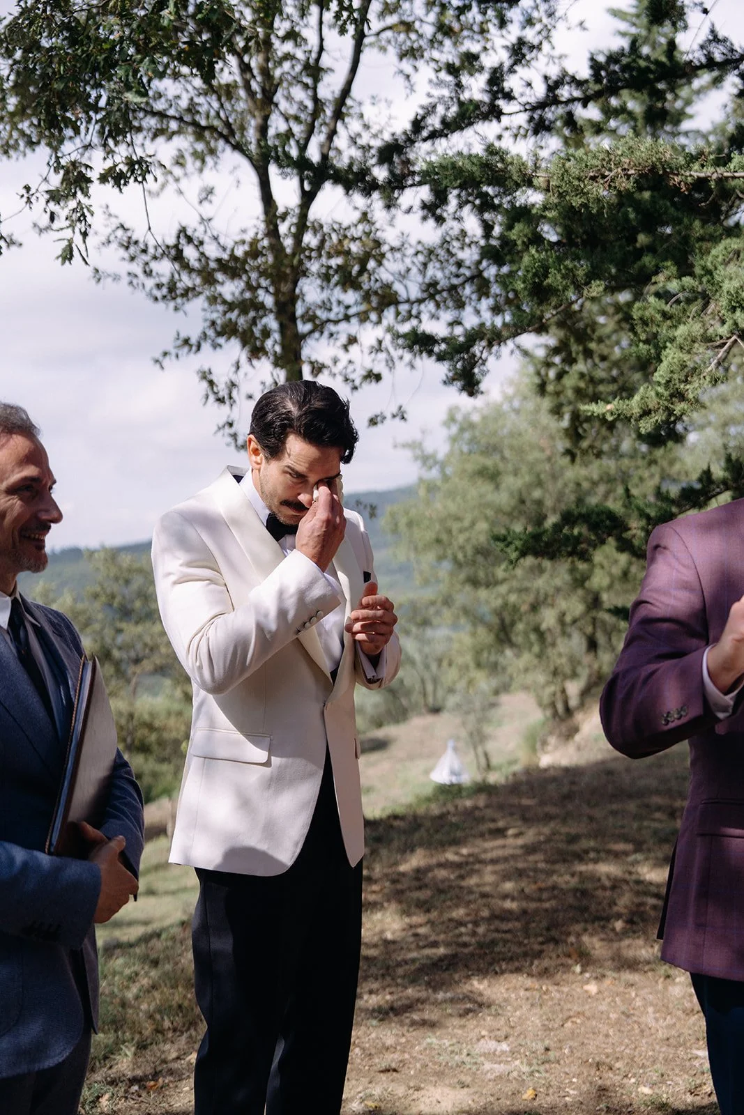 Man in white tuxedo with black bowtie wiping his eye while standing outdoors among other men in suits, trees, and cloudy sky in the background.